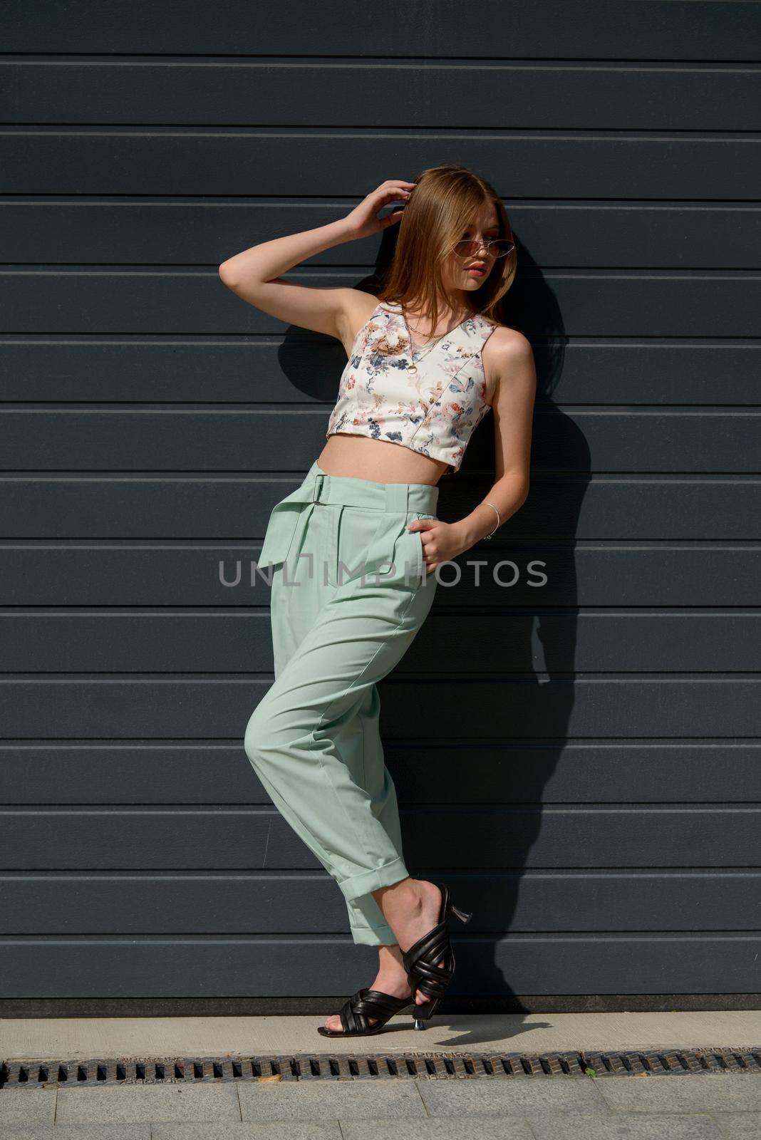 Fashion portrait of young woman wearing sunglasses, top, slingbacks, blue suit. Young beautiful happy model posing near gray metal grid.