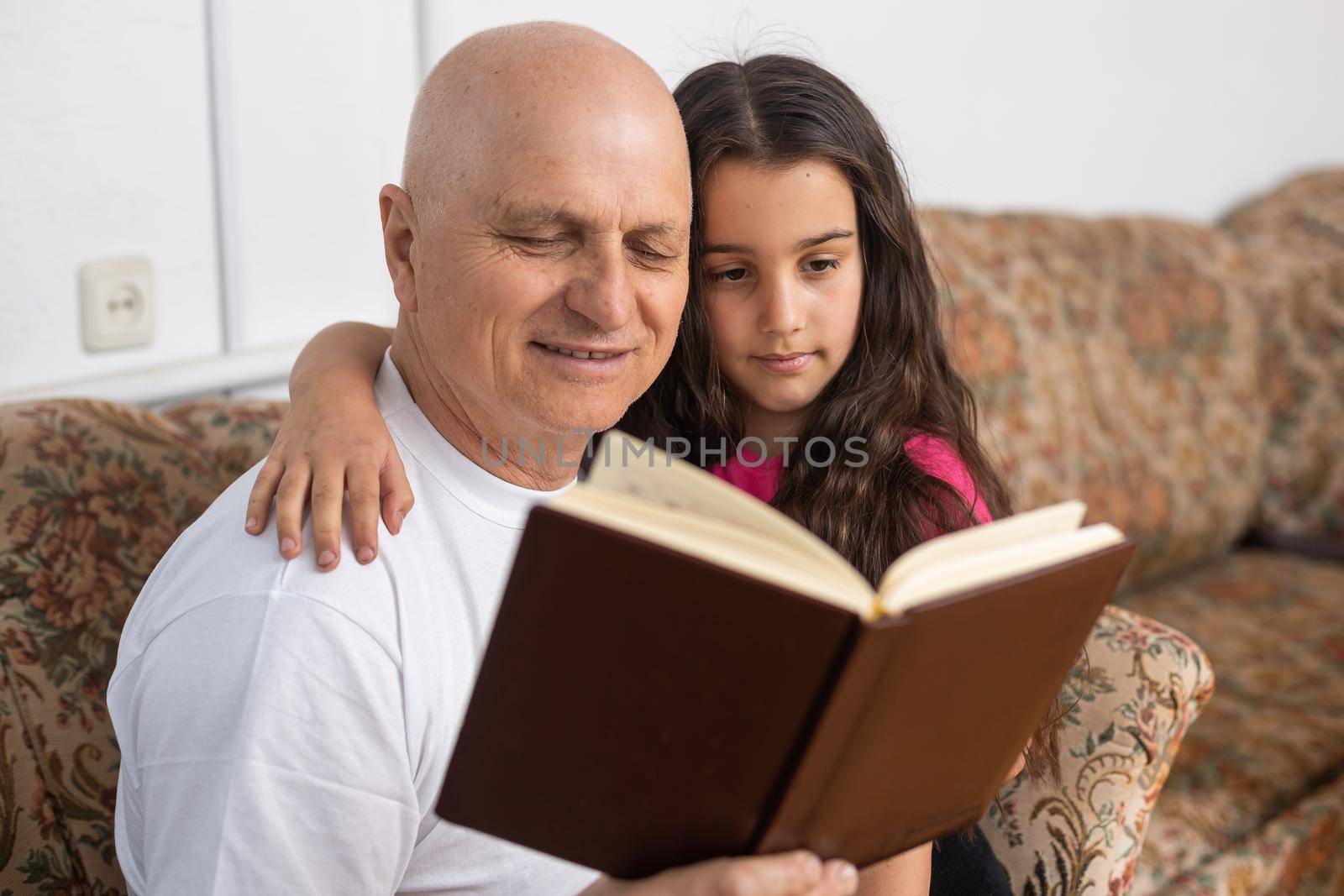 Happy grandfather and granddaughter reading book together at home.
