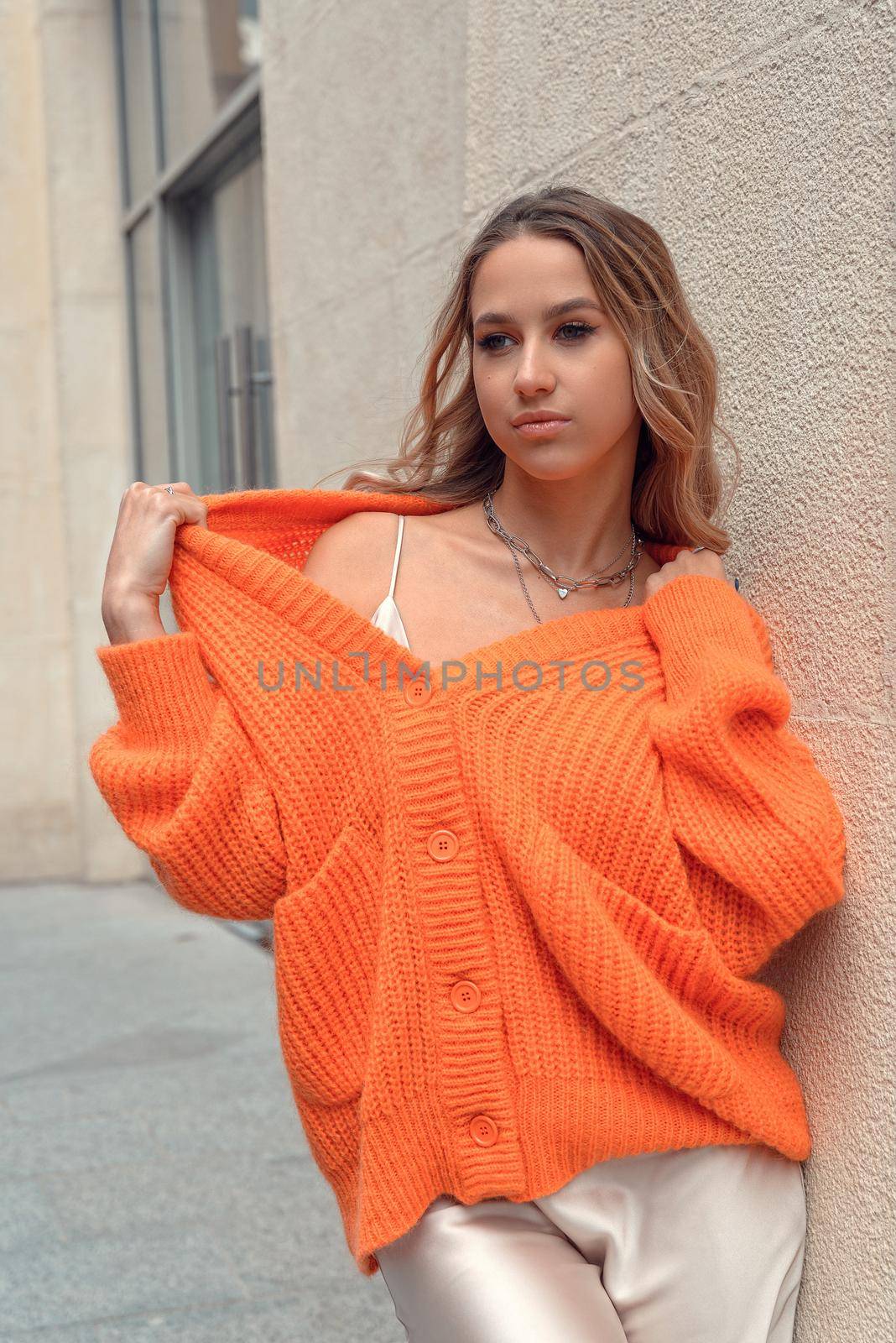 Portrait of fashionable blond women in orange sweater and beige dress posing in the street