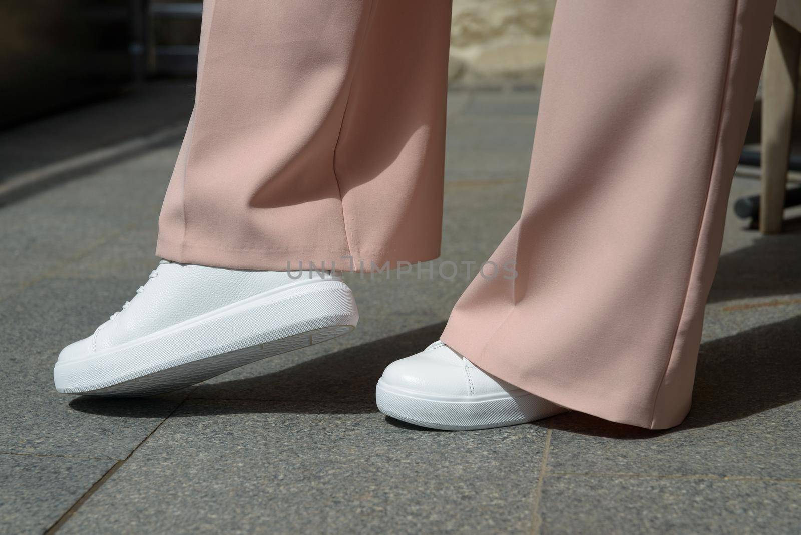 Stylish woman in fashion white sneakers shoes and beige trousers walks around the city. Close-up.