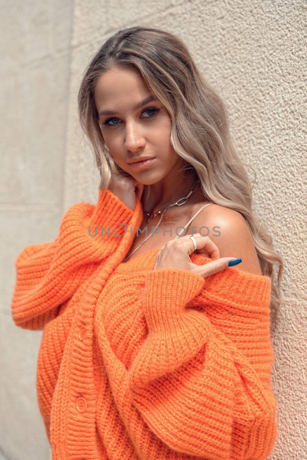 Portrait of fashionable blond women in orange sweater and beige dress posing in the street