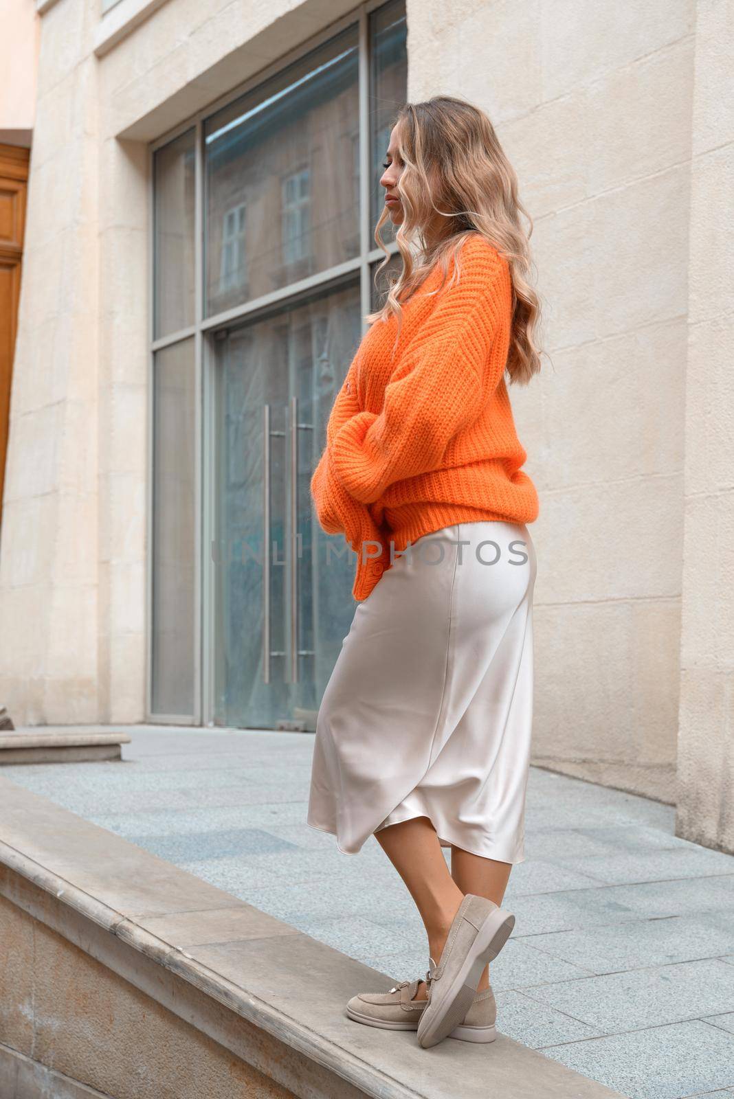 Portrait of fashionable blond women in orange sweater, beige dress and stylish suede loafer shoes posing in the street