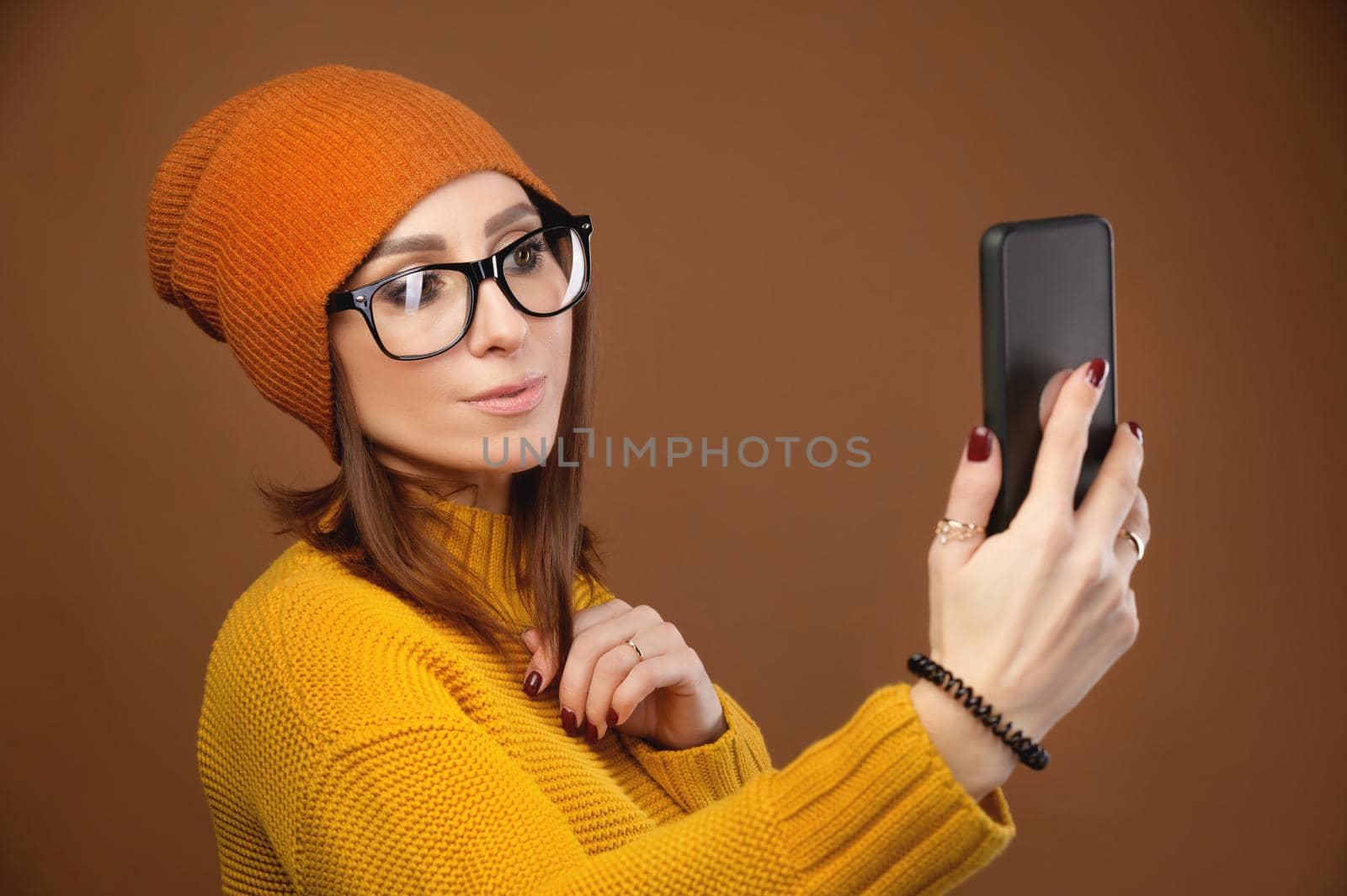 Beautiful young caucasian woman with flowing hair wearing glasses sweater and hat taking a selfie with your smartphone. Studio portrait on a brown background. Copy space.