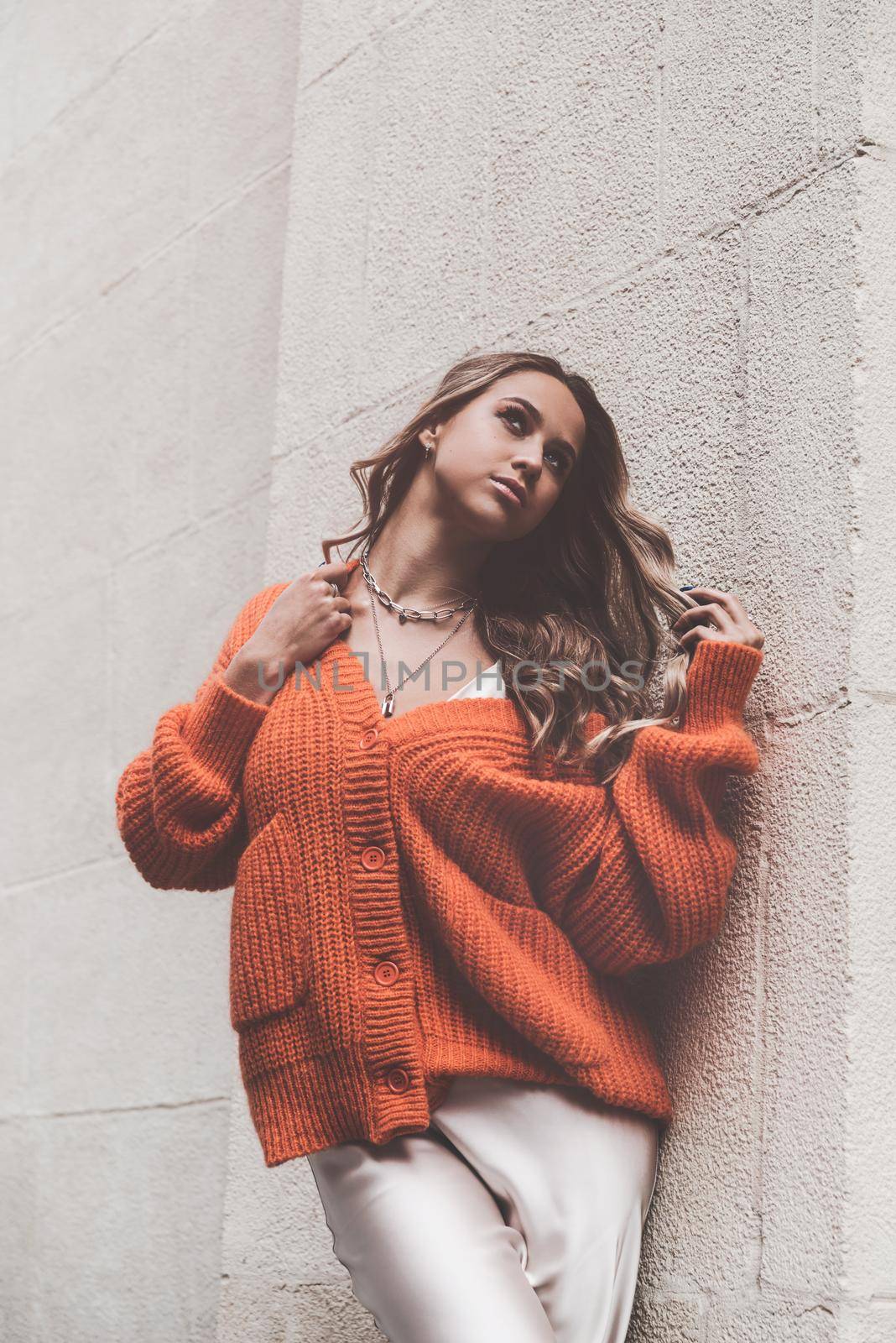 Portrait of fashionable blond women in orange sweater and beige dress posing in the street