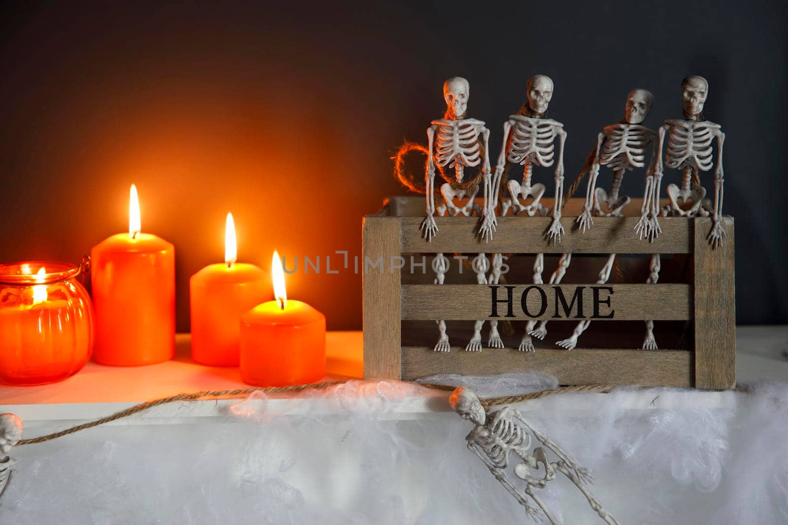 Halloween home decoration. Plastic toy skeletons in wooden box on fireplace against a dark blue wall. A garland of skeletons. Cobweb on the dresser. Orange candles and lantern.