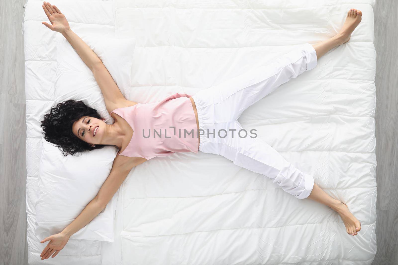 Top view of happy woman laying like star on white bedding, wake up well rested in morning, king size bed, good relaxation. Chilling on bed in pyjamas at home. Comfort, sleep, rest concept