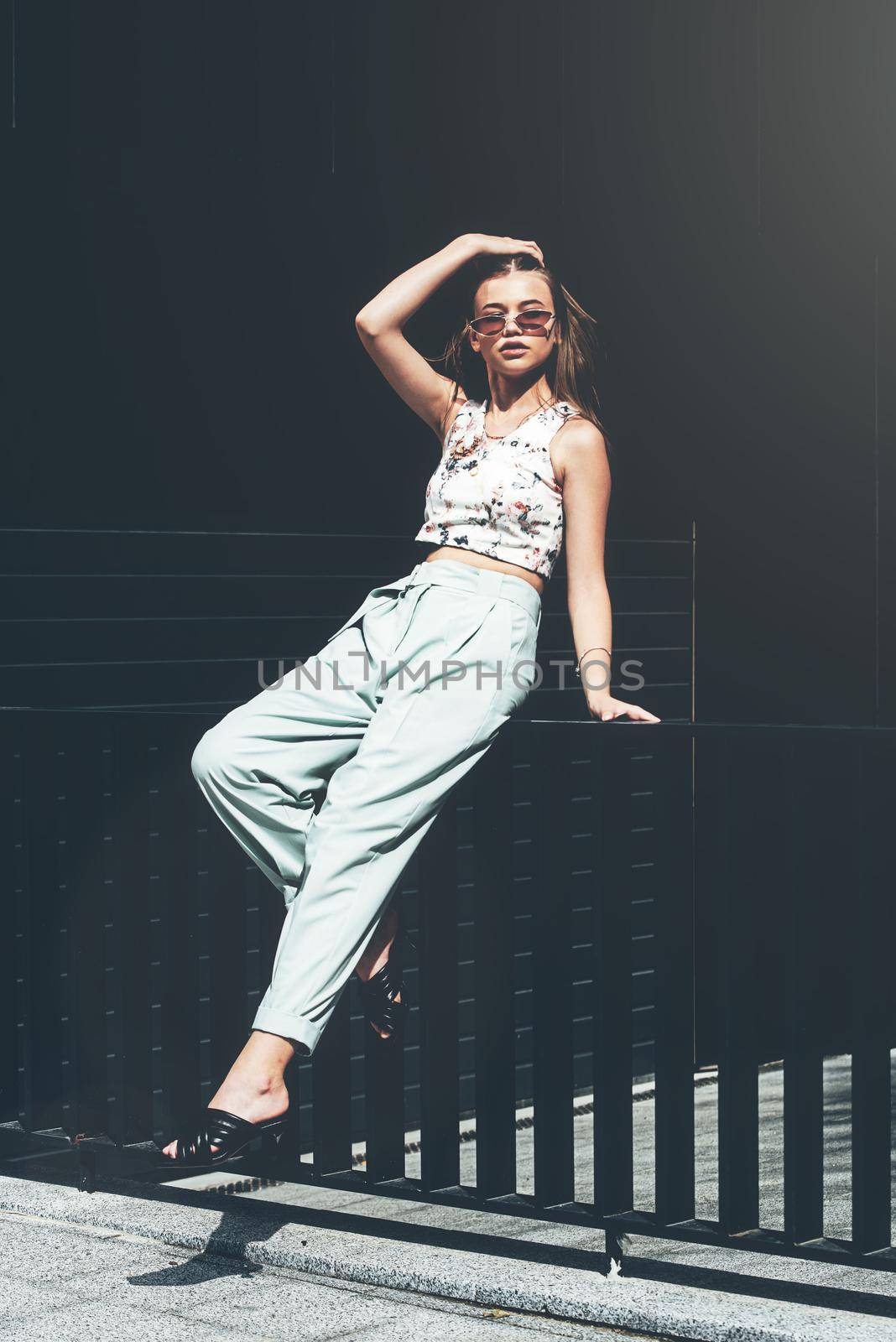 Fashion portrait of young woman wearing sunglasses, top, slingbacks, blue suit. Young beautiful happy model posing near gray metal grid by Ashtray25