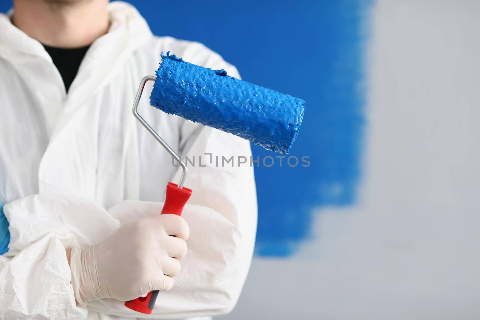 Close-up of painter man hand holding roller tool for painting walls, blue colour on equipment, worker in uniform. Copy space in right. Renovation, interior design, redecoration concept