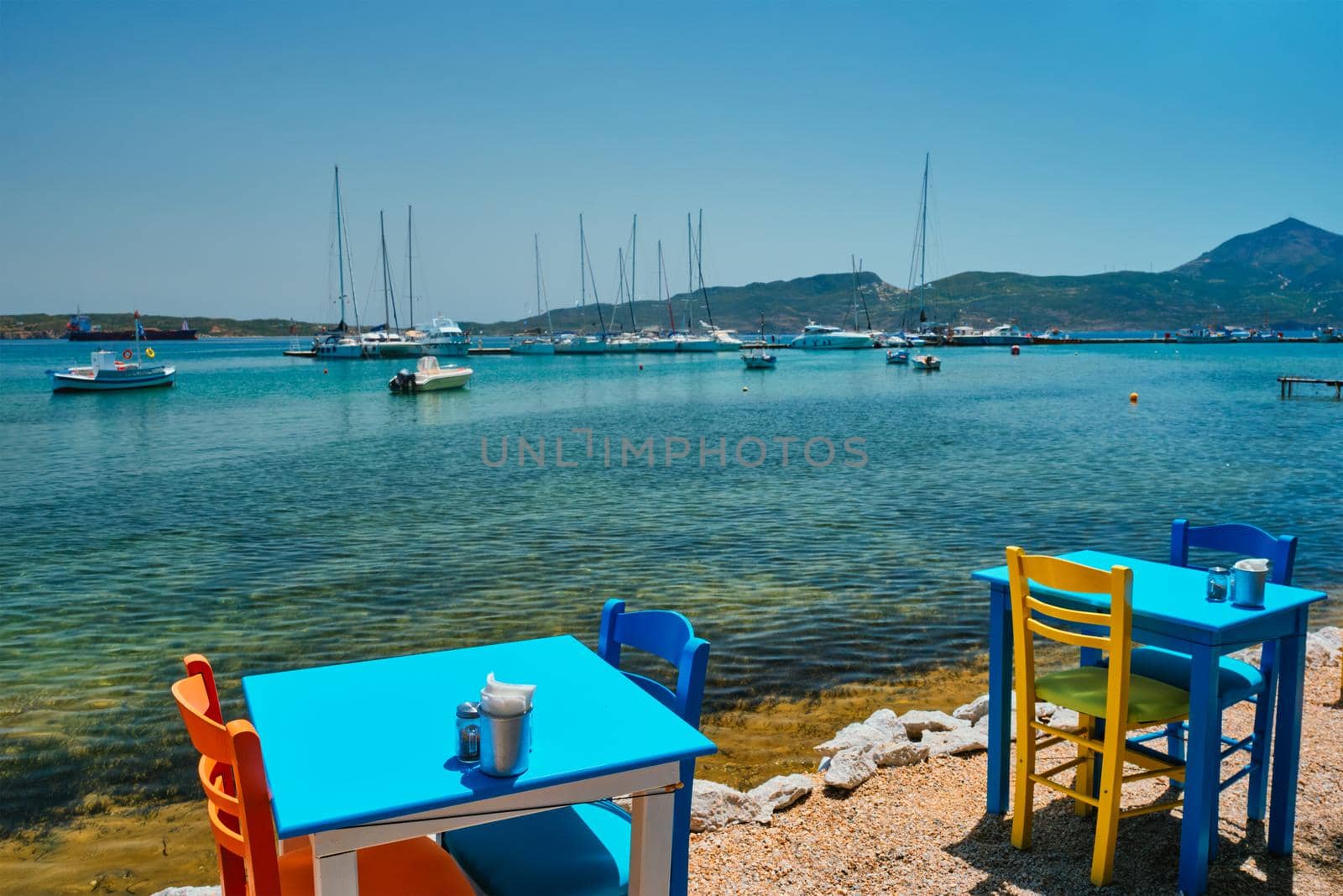 Cafe tableon beach in Adamantas town on Milos island with Aegean sea with boats in background by dimol