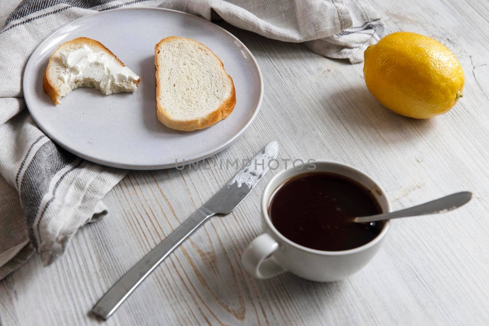 A cup of coffee, two pieces of sugar, a lemon, a piece of bread with curd cheese spread on it on a white ceramic plate with a knife on the table. by elenarostunova
