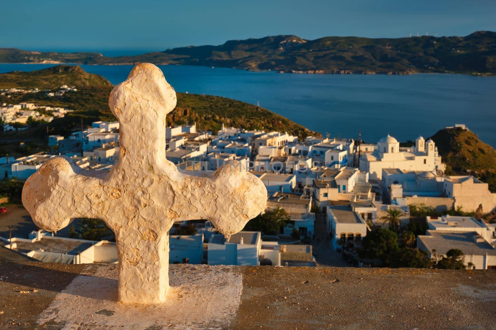 Greek Orthodox Christianity concept background - Christian cross with Plaka village on Milos island in background on sunset. Plaka town, Milos island, Greece. Focus on flowers