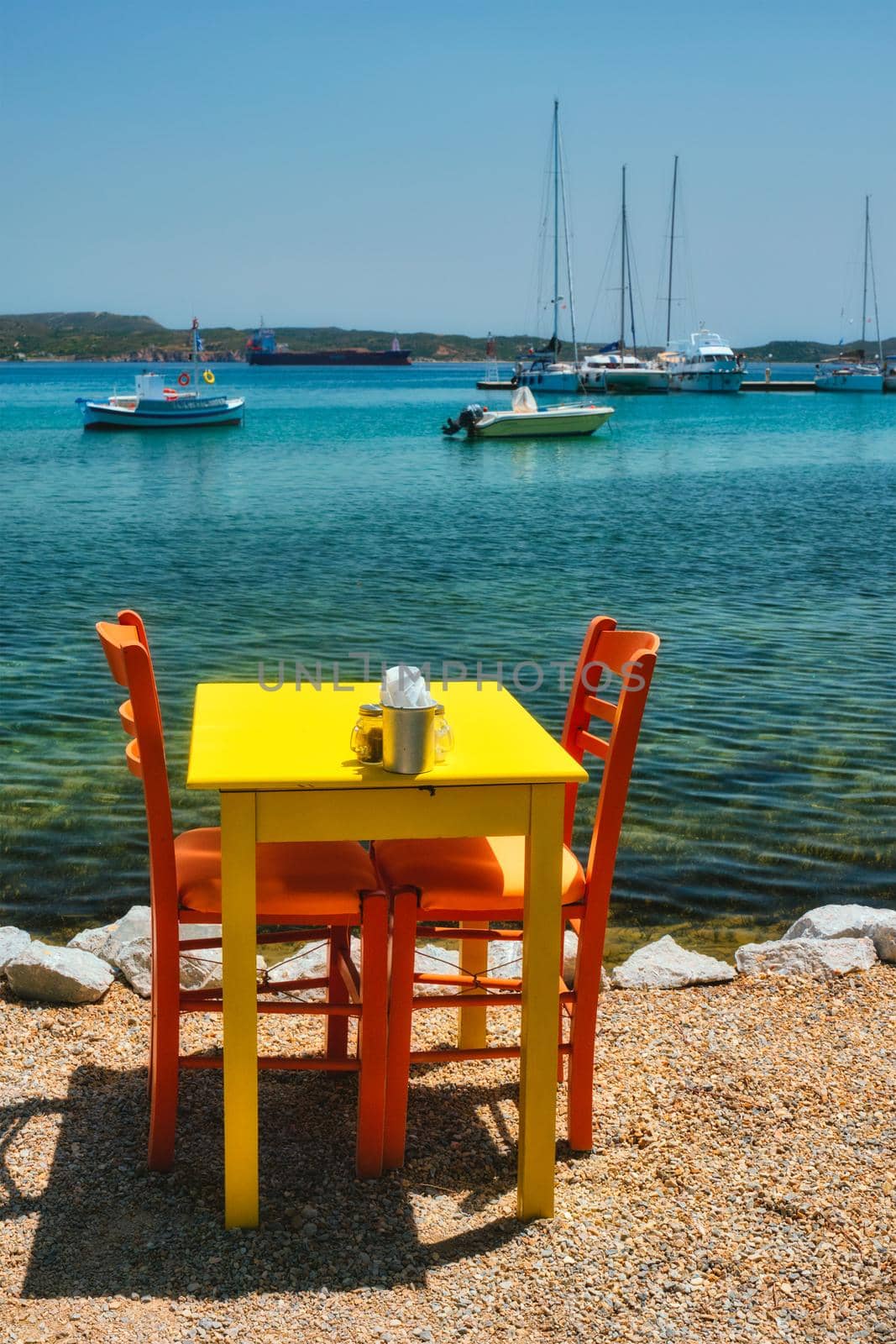 Cafe tableon beach in Adamantas town on Milos island with Aegean sea with boats in background by dimol