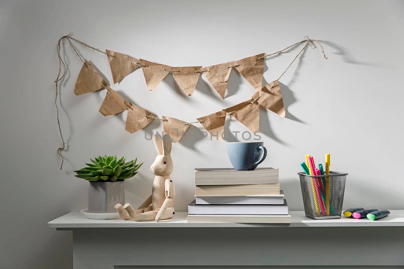 Echeveria in geometric pot, stack of books, pens in a pencil holder, felt-tip pens, wooden figurine of a hare are on shelf. Garland of craft paper flags on the wall above the shelf. Place for text by elenarostunova