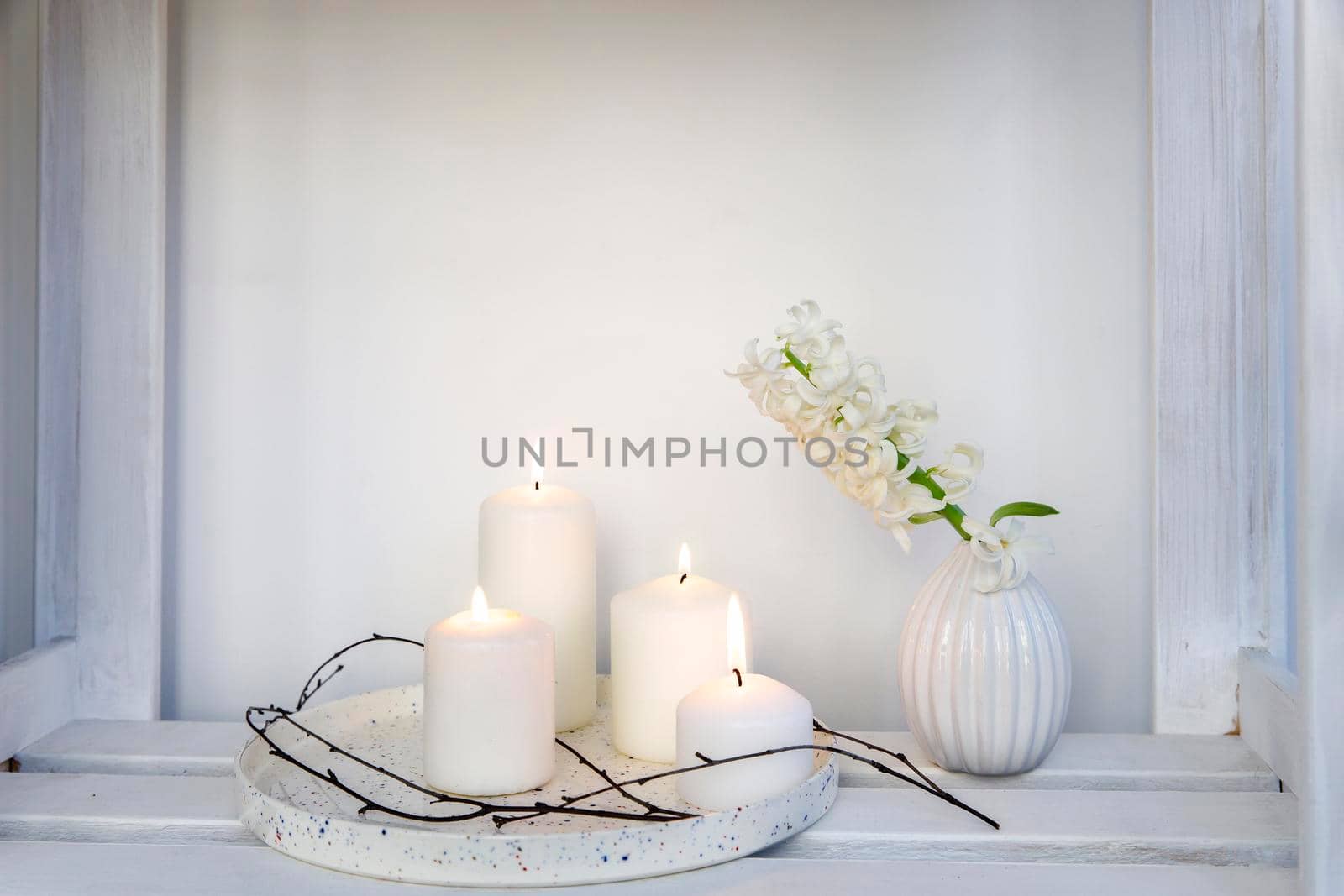 A bouquet of white cut hyacinth a in a small white corrugated vase and three large burning candles on a round tray are on a beige table. Place for text