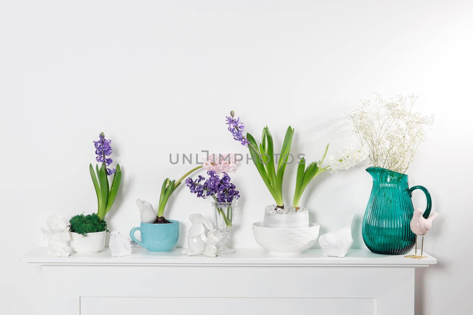 White hyacinth in a large porcelain bowl, figurines of hares and a bird, are on the fireplace against the white wall. Layout. Spring concept by elenarostunova
