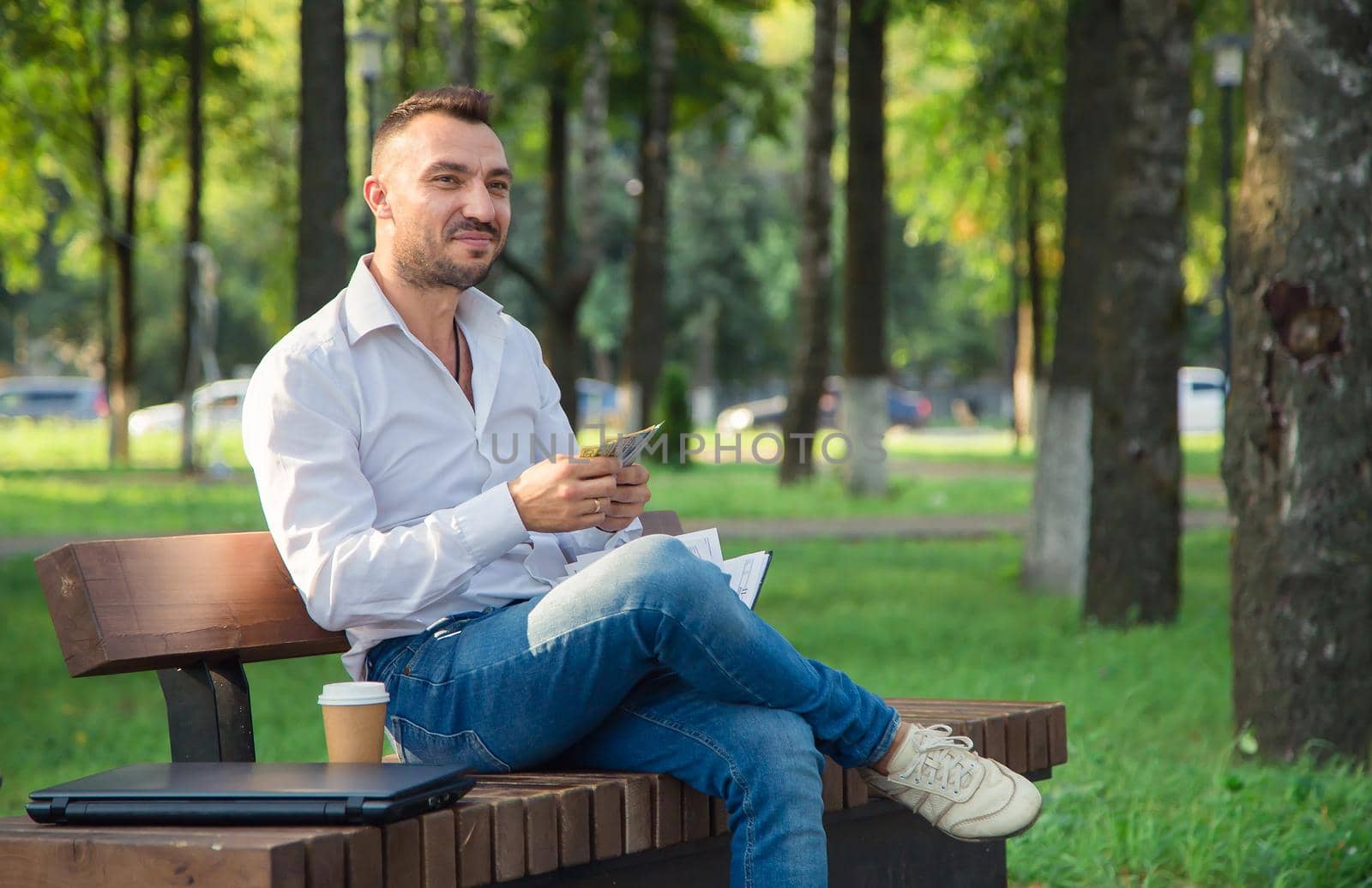 A smiling man is sitting on a bench in the park, counting dollars. A young man on a background of green trees, a hot sunny summer day. Warm soft light, close-up.