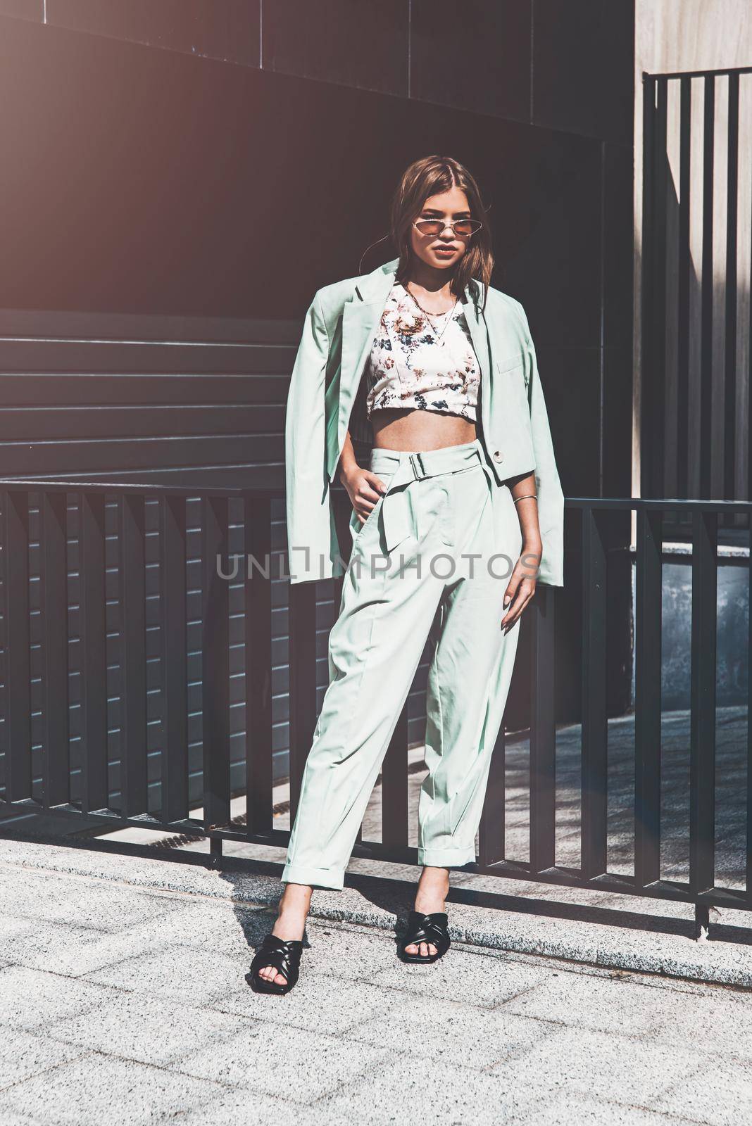 Fashion portrait of young woman wearing sunglasses, top, slingbacks, blue suit. Young beautiful happy model posing near gray metal grid.