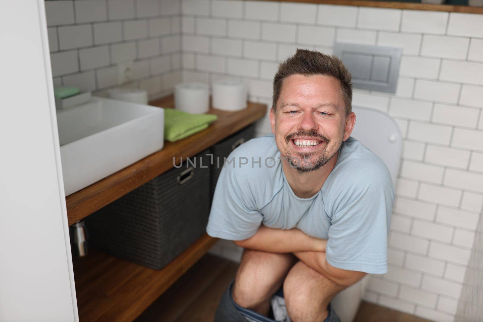 Portrait of man feeling relief after using toilet, fulfill natural need. Man in bathroom with modern interior, white tiles, wooden commode, new sink and decor. Wc, renovation, nature calls concept