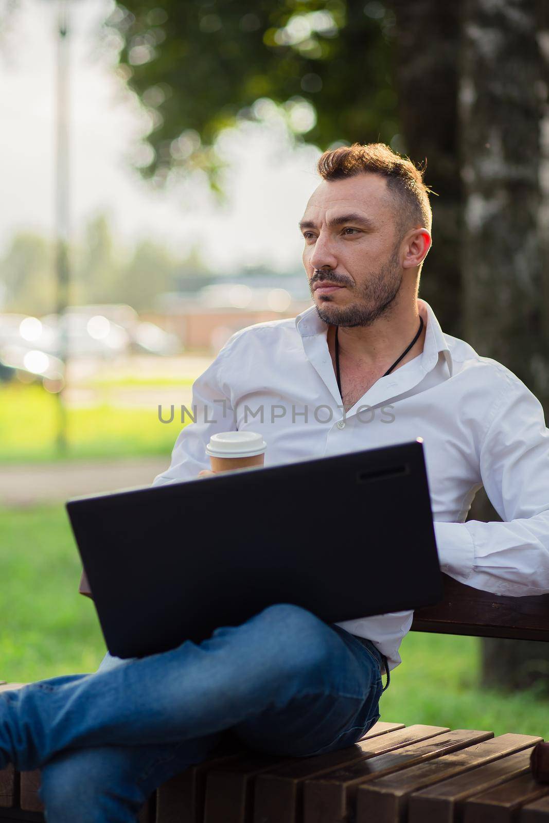 Dreamy man works in the park with a laptop, drinks coffee. A young man on a background of green trees, a hot sunny summer day. Warm soft light, close-up.