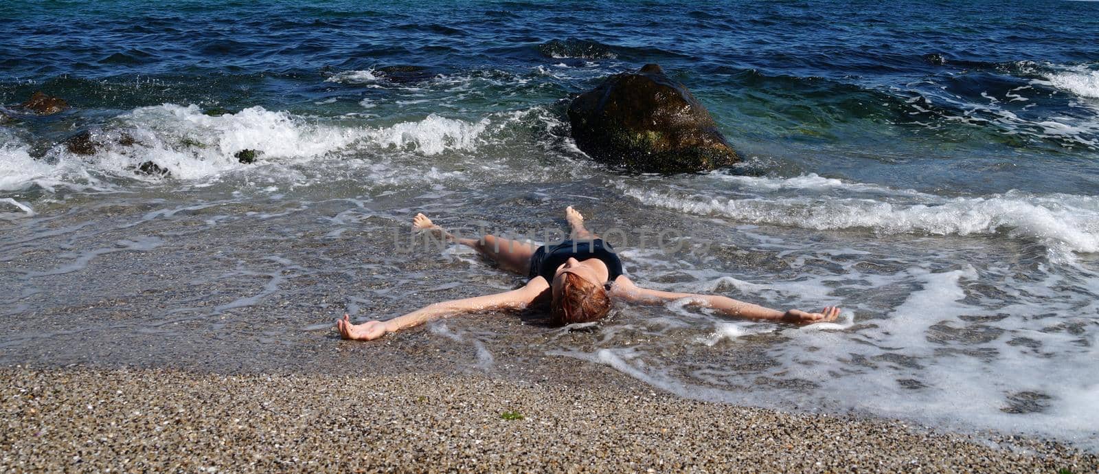 a girl in a black swimsuit lies with her arms outstretched on the shore in sea water.