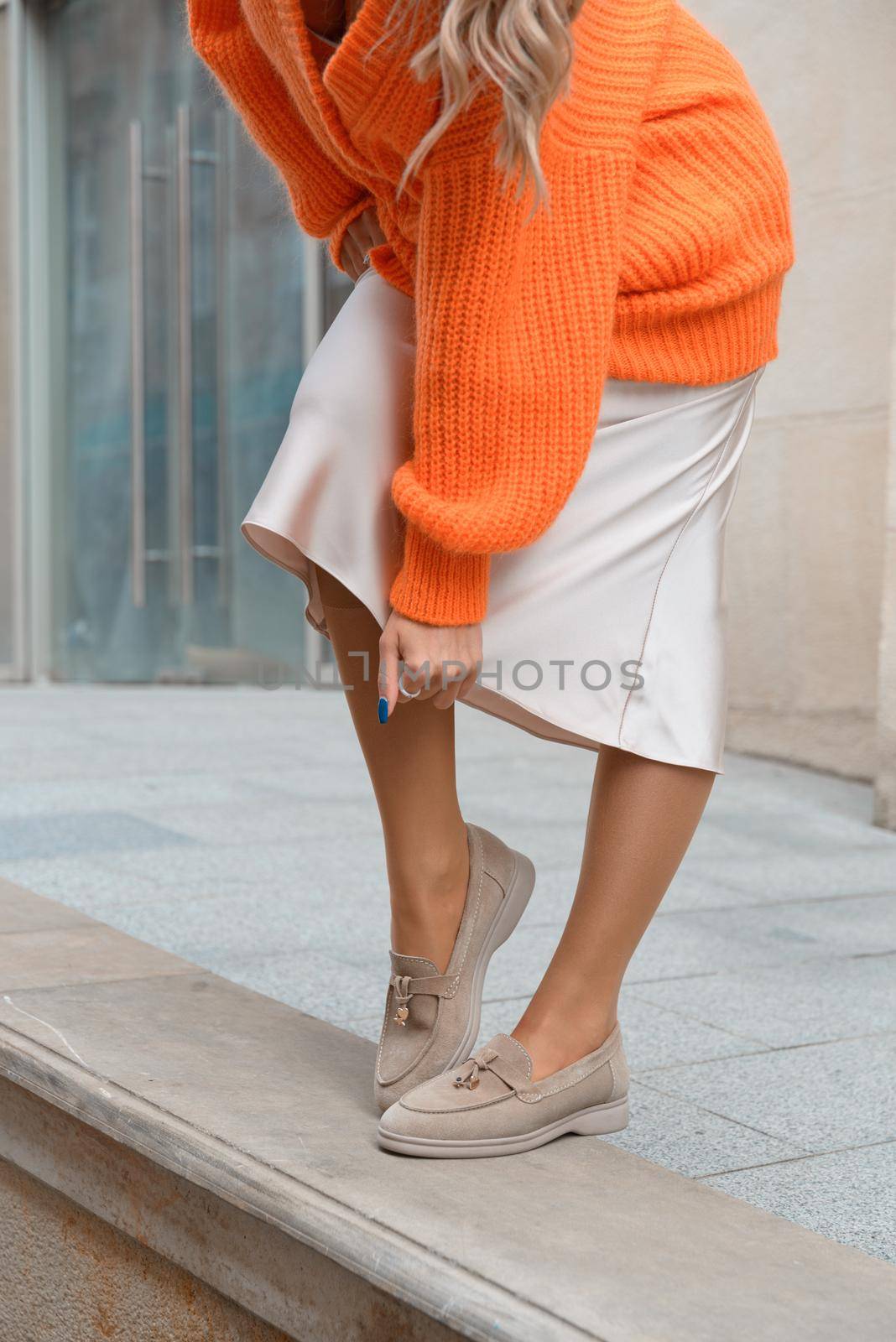 Portrait of fashionable blond women in orange sweater, beige dress and stylish suede loafer shoes posing in the street