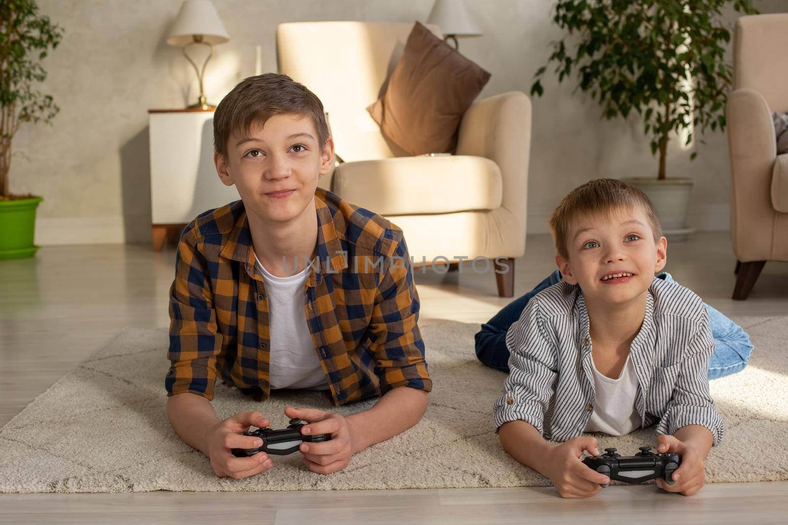 Two joyful boys lie on the floor in a room playing video games with joysticks. Close up