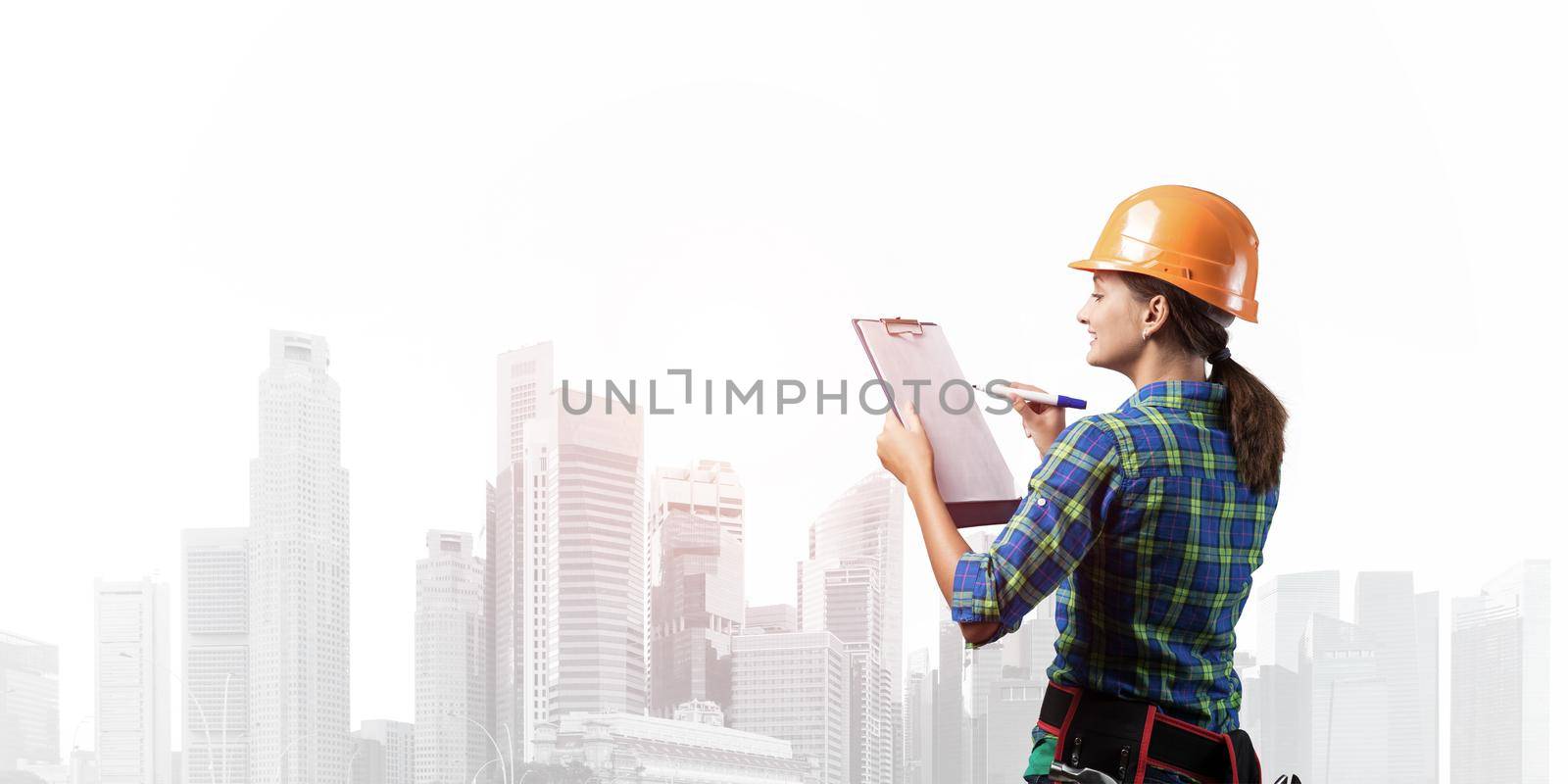 Young building controller with clipboard standing and filling examination report. Side view of woman construction specialist in workwear and hardhat writing in checklist. Professional house audit
