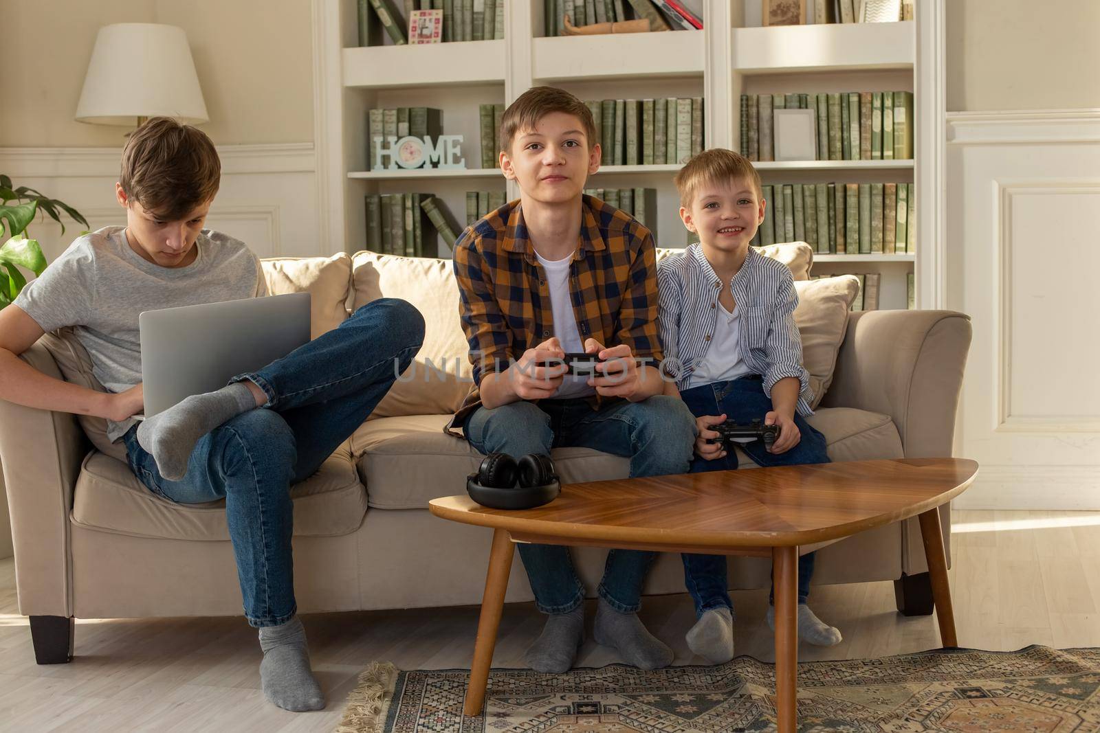 Three brother boys playing computer games on a laptop and video games with joysticks in their hands by Zakharova