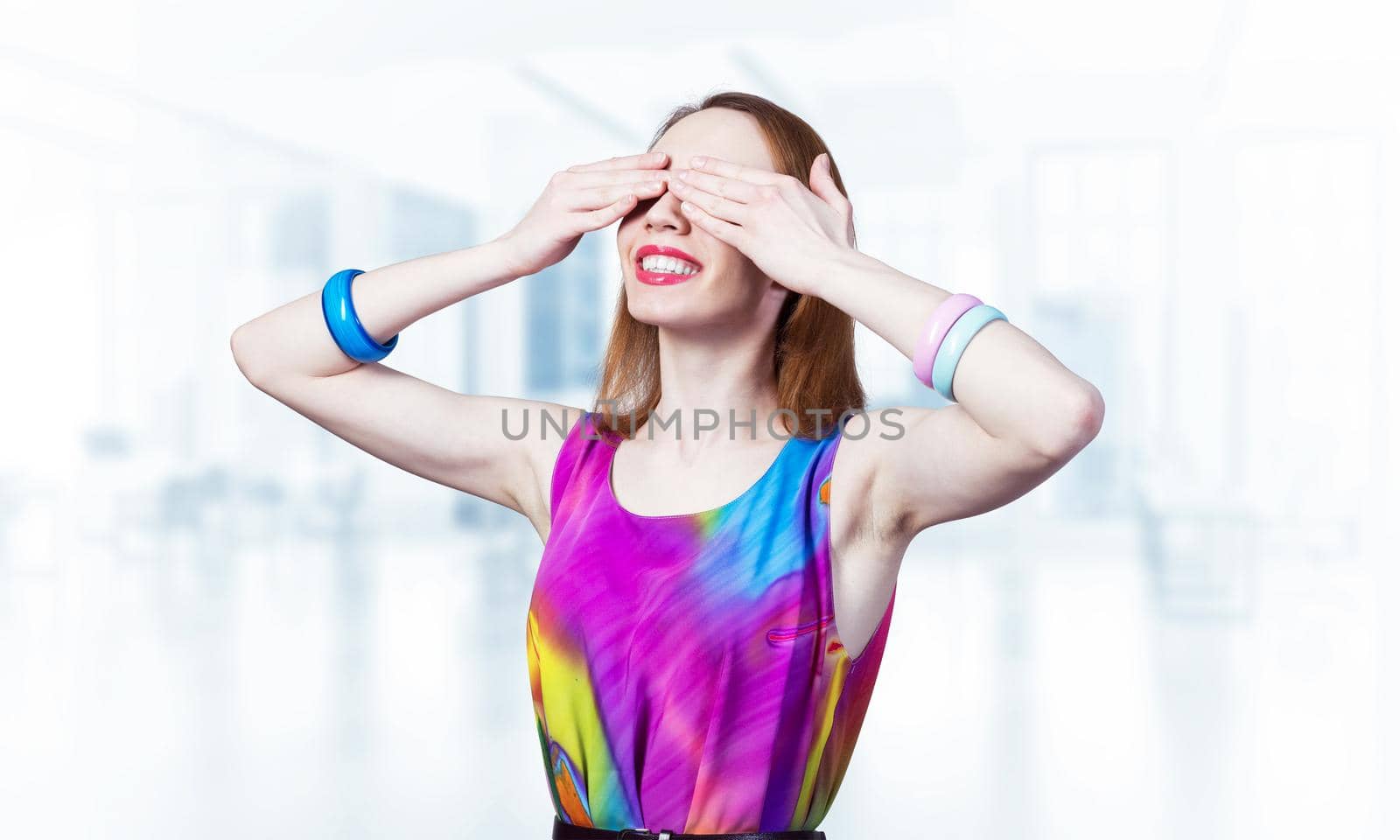 Redhead beautiful girl closing her eyes with hands. Charming lady wears bright dress and bracelets standing on blurred office interior background. Portrait of young woman waiting for pleasant surprise