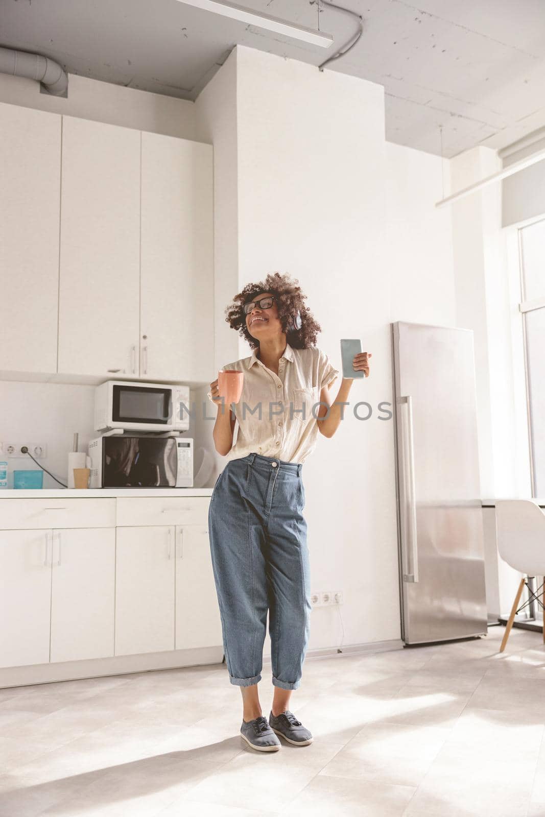 Full-length photo of cheerful African American female using wireless headphones and dancing on the kitchen while holding phone