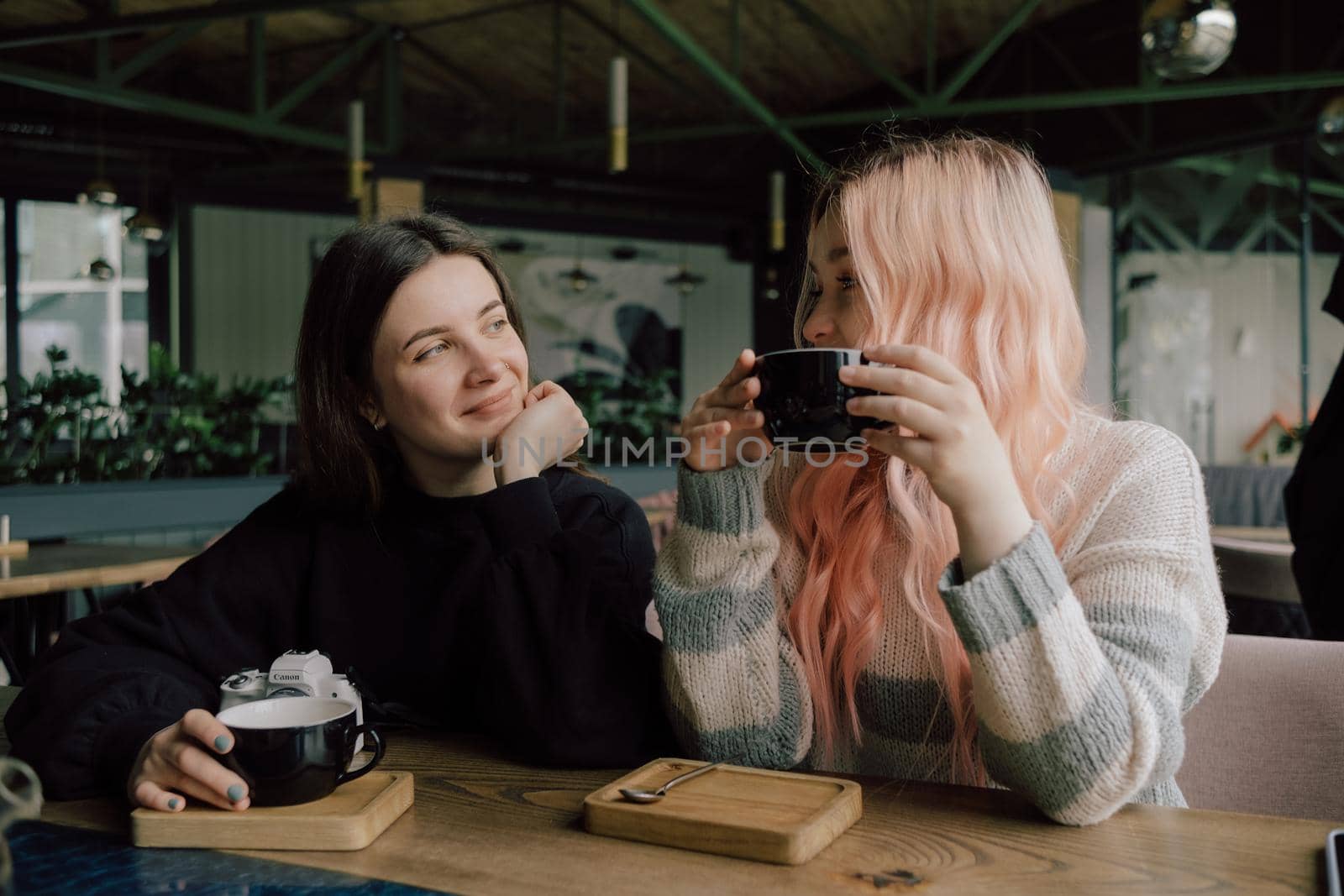 Interracial lesbian couple each other with shy smile, holding hands during lunch at restaurant. Happy redhead woman confessing love to her stylish Asian American girlfriend with pink hair hairstyle