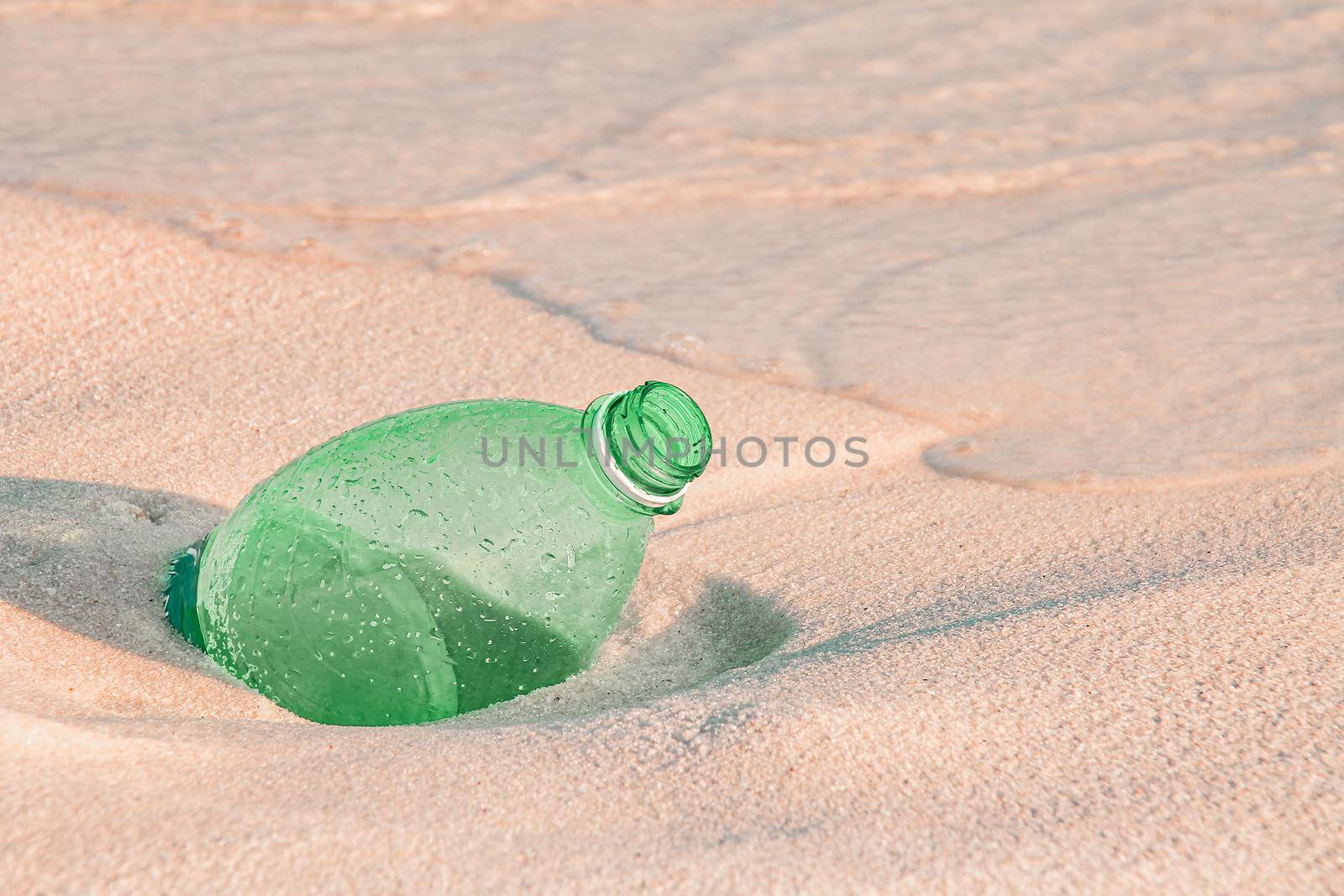 Close up of a green plastic water bottle buried in the sandy beach