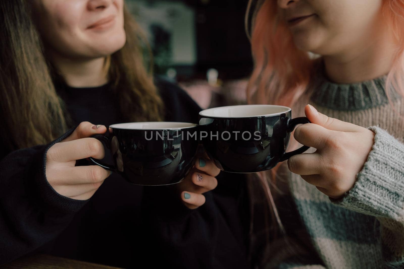 Interracial lesbian couple each other with shy smile, holding hands during lunch at restaurant. Happy redhead woman confessing love to her stylish Asian American girlfriend with pink hair hairstyle. closeup. selective focus on cups