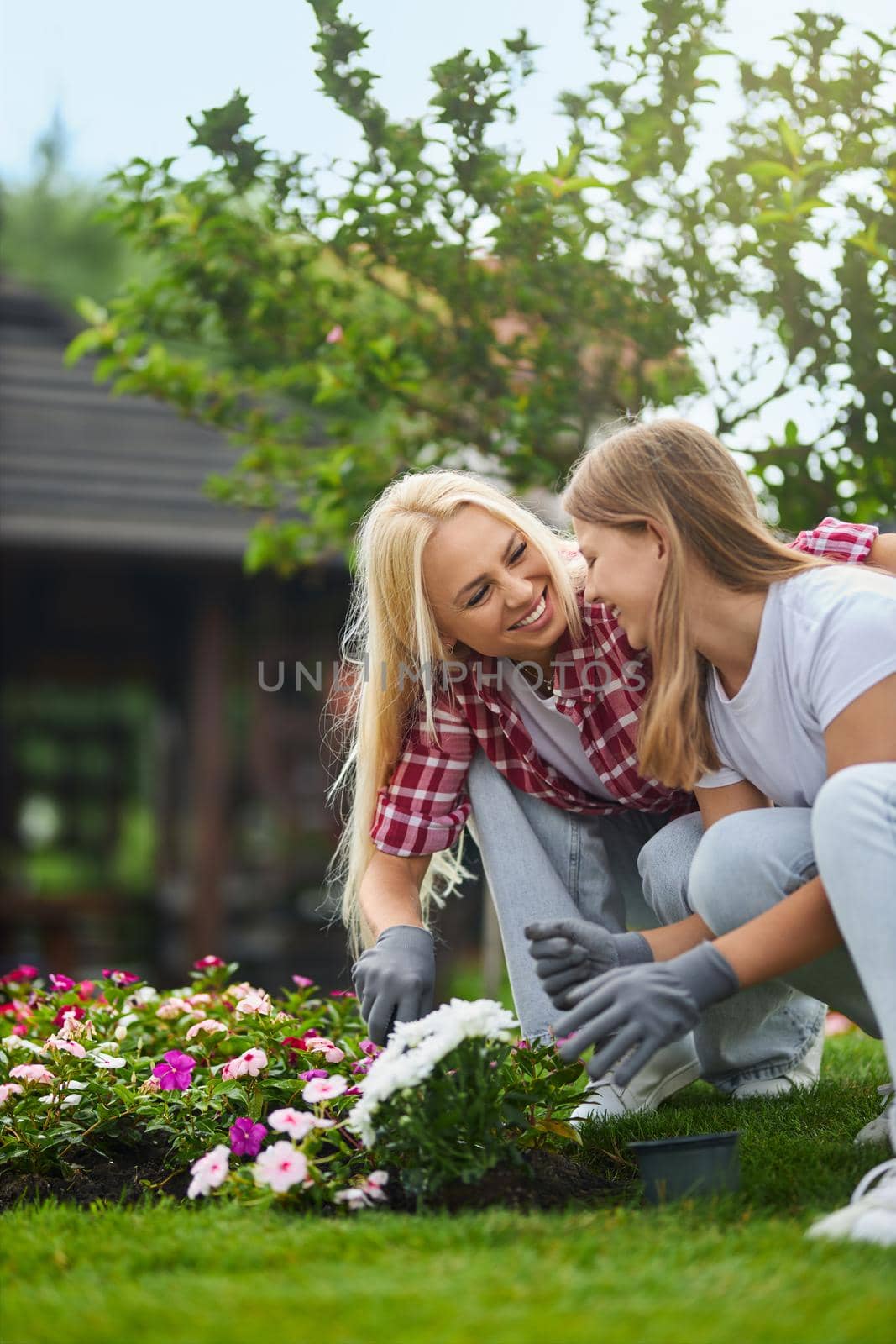 Caring caucasian mother teaching her teen daughter to take care of plants on backyard. Woman and girl squatting on flowerbed, laughing and hugging during working process.