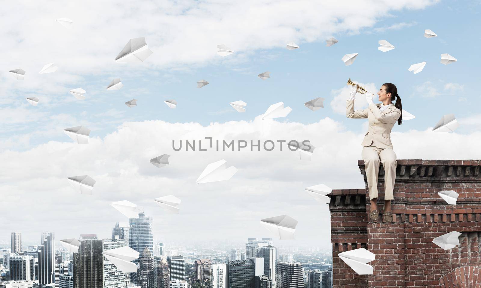 Beautiful young woman with trumpet on roof. Brave girl in white business suit with music brass instrument sitting on edge of roof. Flying paper planes in blue sky. Female musician playing music