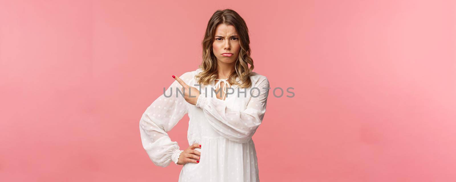Portrait of judgemental upset cute blond girl in white dress, sulking and frowning with blame, accuse someone make offensive thing, pointing finger upper left corner insulted and gloomy.