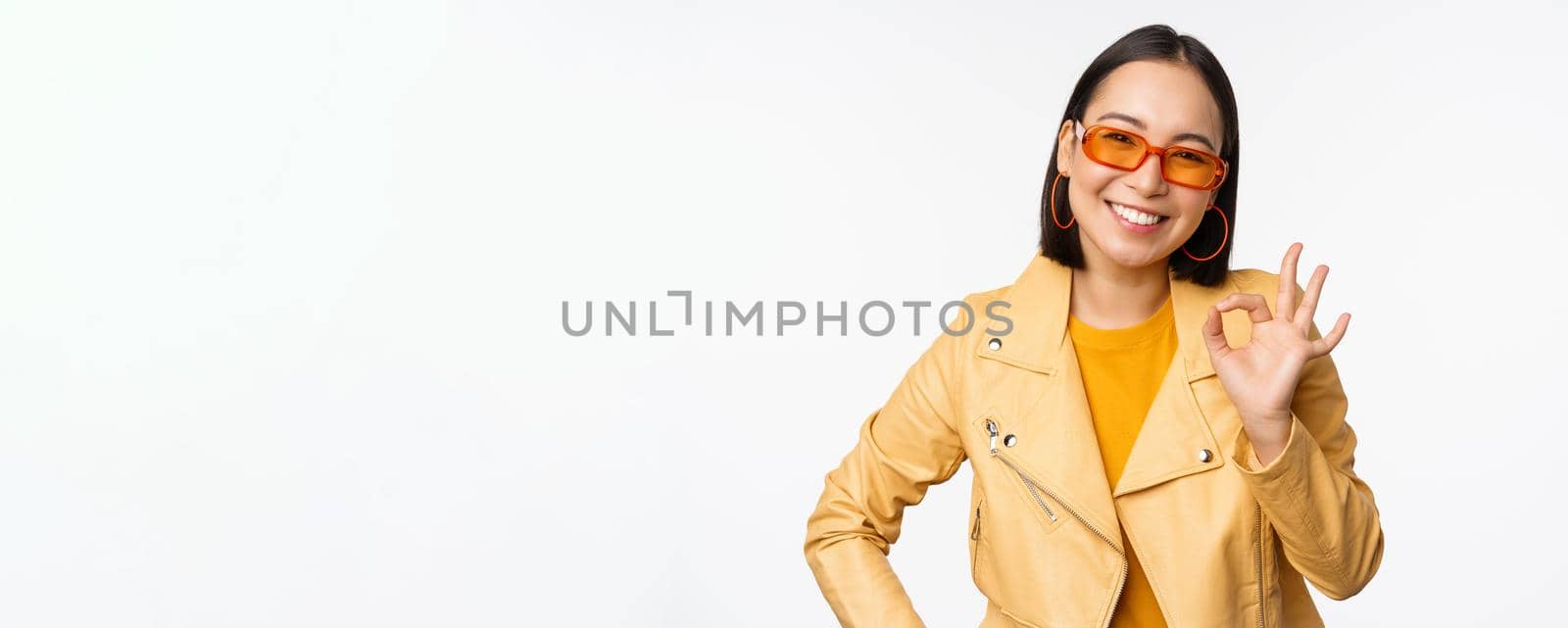 Excellent. Smiling asian girl shows okay, ok sign, nod in approval, recommending smth good, standing over white background by Benzoix