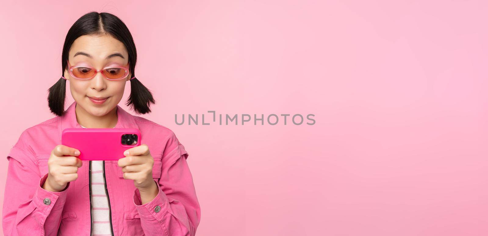 Portrait of happy asian girl playing on smartphone, watching videos on mobile phone app, standing over pink background by Benzoix