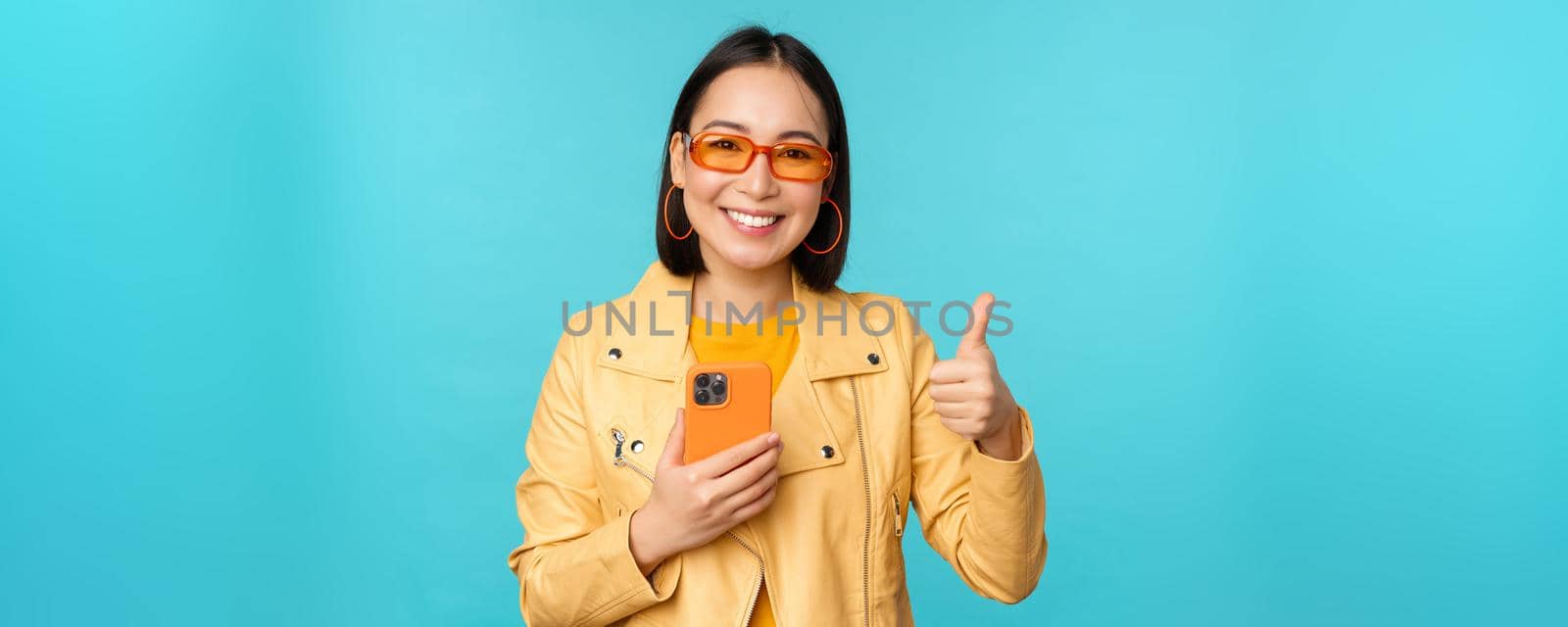 Smiling asian woman showing thumbs up, recording on mobile phone, using smartphone app and recommending it, standing over blue background by Benzoix