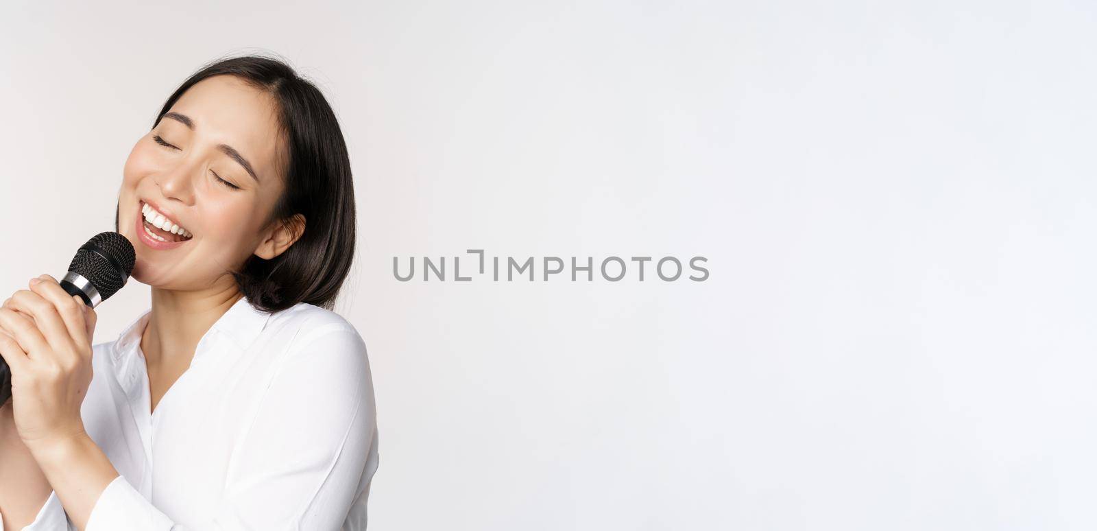 Close up portrait of asian woman singing in microphone at karaoke, standing over white background.