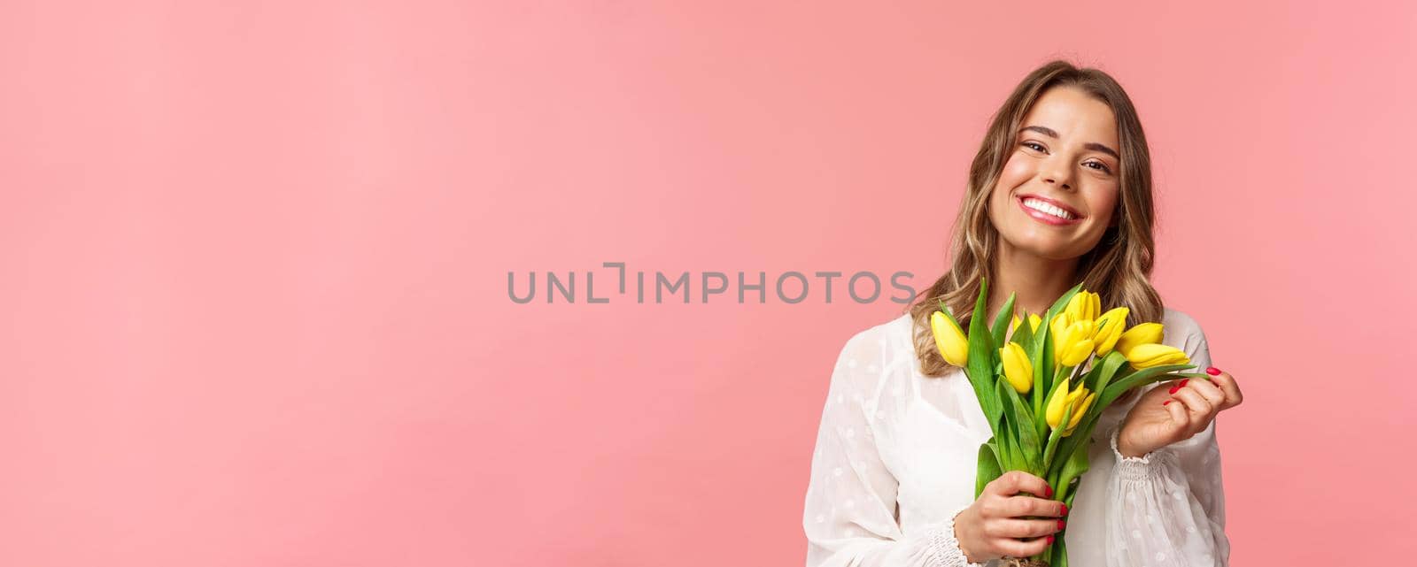 Spring, happiness and celebration concept. Close-up of lovely happy young blond girl in white dress, daydreaming after perfect date, holding yellow tulips and smiling at camera, pink background.