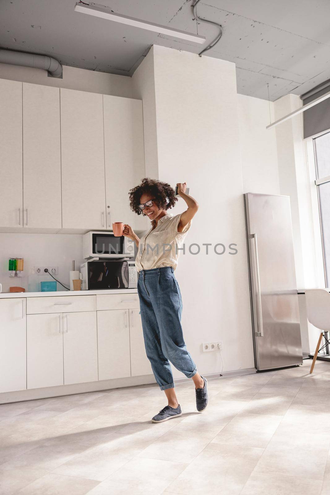 Full-length photo of African American woman holding cup of hot drink while using headphones and dancing on the kitchen