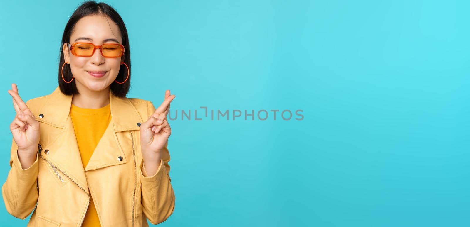 Smiling beautiful asian woman wishing, cross fingers for good luck and looking hopeful, standing over blue background. Copy space