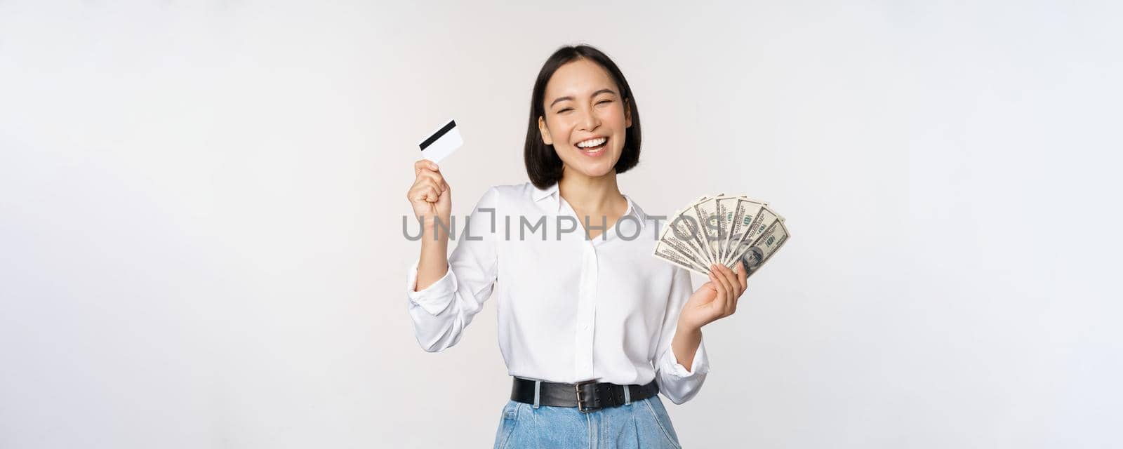 Happy korean woman holding credit card and money dollars, smiling and laughing, posing against white studio background.