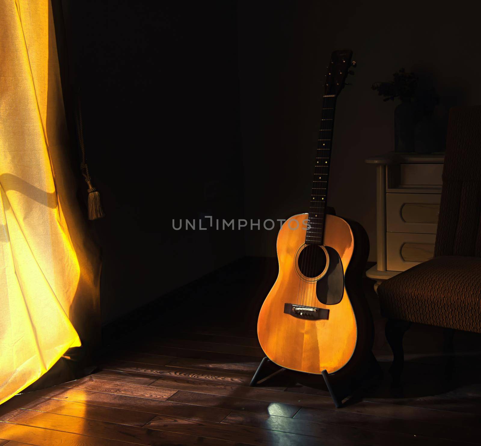 Acoustic Spanish guitar on a stand in the moody shadows of a dark room with bright light coming in from behind a curtain