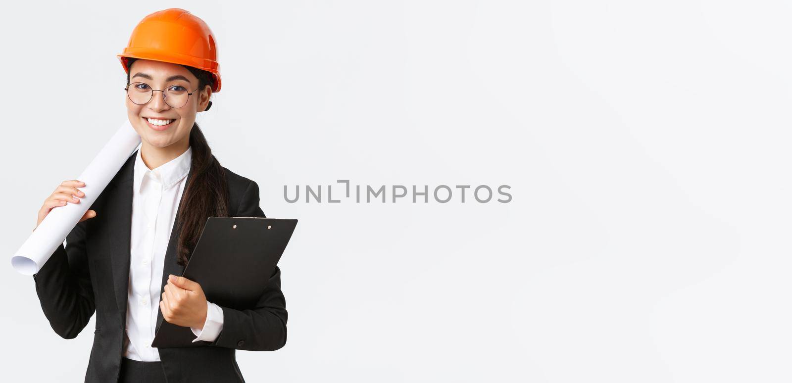 Successful asian female architect in business suit and helmet holding blueprints and clipboard with notes, inspector looking at construction works, smiling at camera, white background.