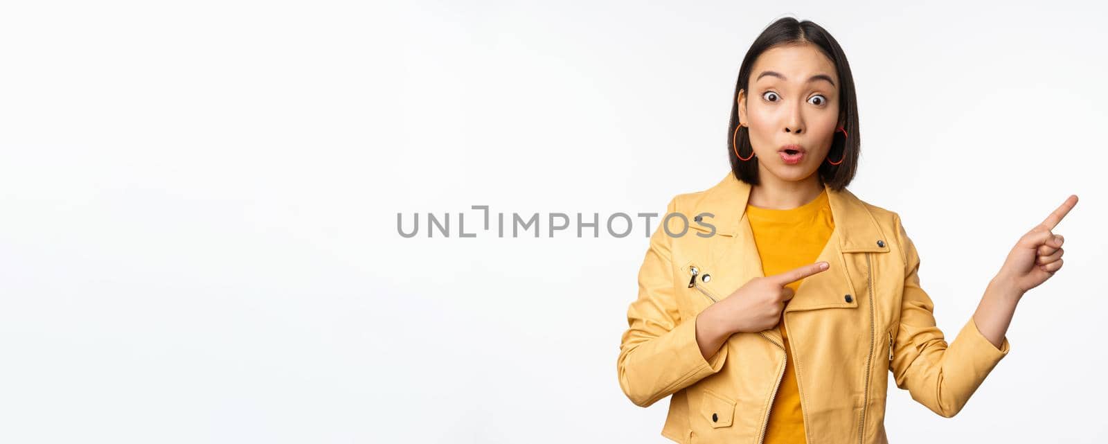 Enthusiastic asian girl pointing fingers right, showing advertisement and smiling, inviting to the store, standing over white background by Benzoix