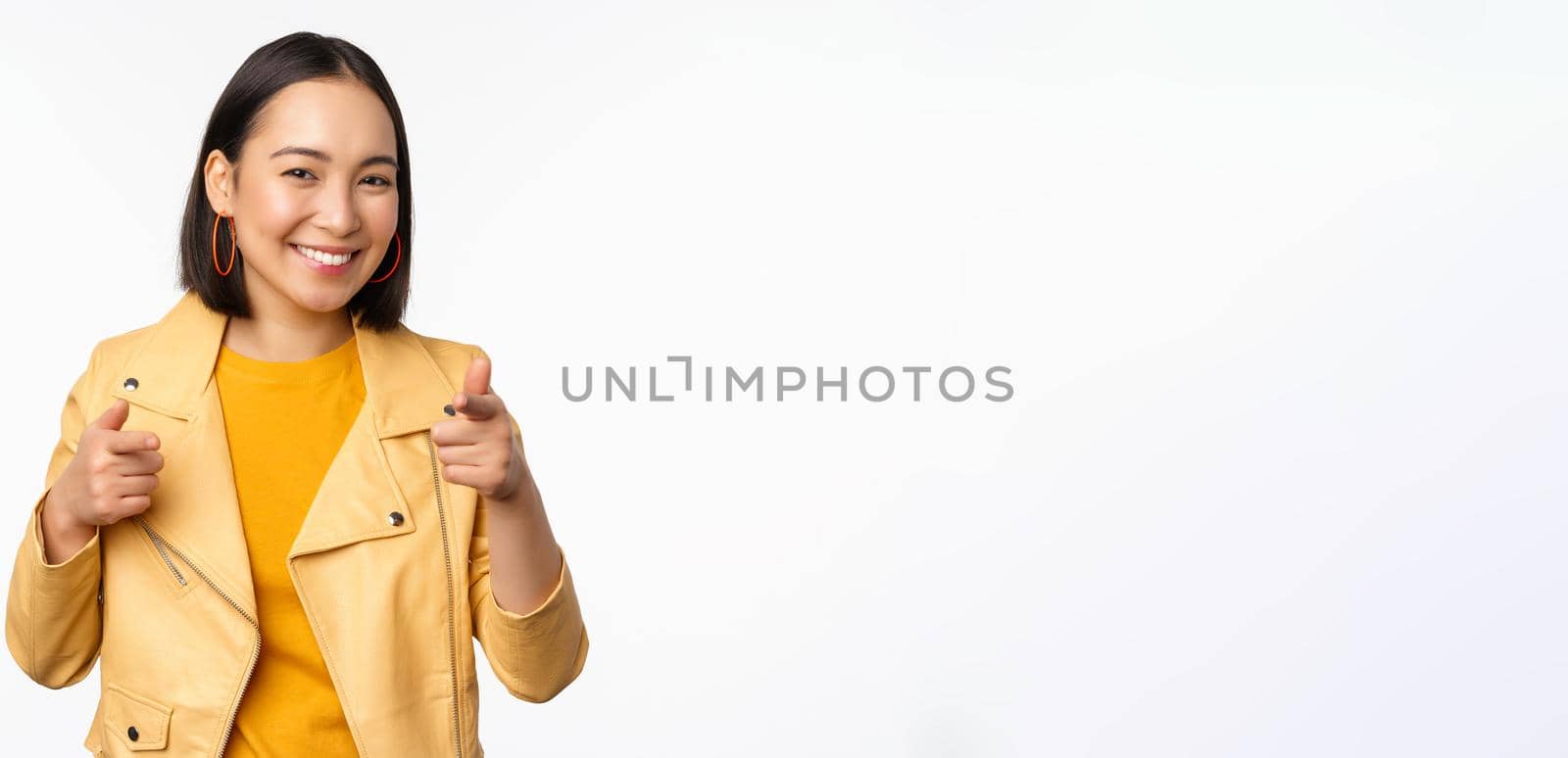 Hey you. Happy smiling asian woman pointing fingers at camera, choosing, congratulating and praising you, standing against white background.