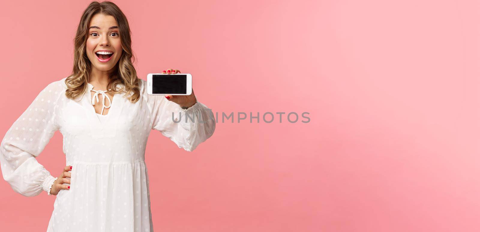 Portrait of cheerful, upbeat attractive blond caucasian girl in white dress, showing smartphone display, hold mobile phone horizontal smiling camera amazed, stand pink background by Benzoix