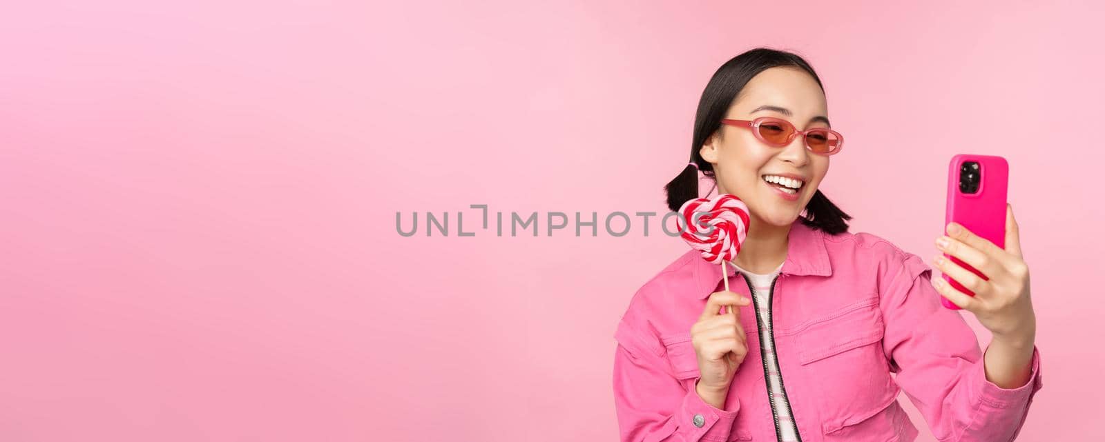 Portrait of stylish, happy asian girl taking selfie with candy, lolipop sweets and smiling, taking photo with mobile app, standing over pink background.